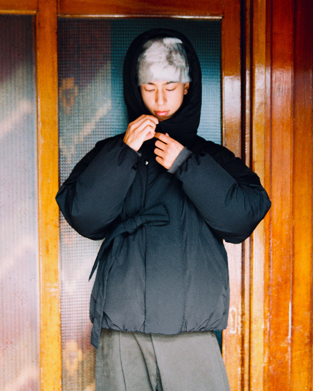 A person wearing the BLACK IBUL DOWN PUFFER by Sundae School, an oversized down jacket with a detachable hood, and a gray furry hat stands in front of wooden doors, adjusting the zipper on their jacket.
