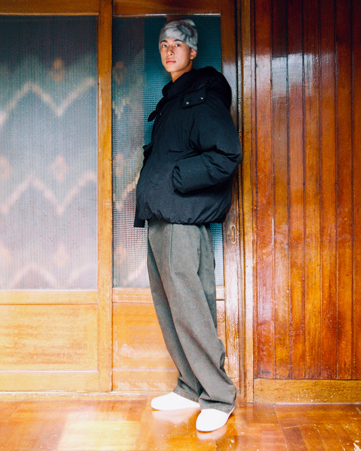 A person stands indoors wearing the BLACK IBUL DOWN PUFFER by Sundae School, an oversized black cocoon-style down jacket, paired with gray pants, white shoes, and a gray fuzzy hat. The background features wooden doors and panels.