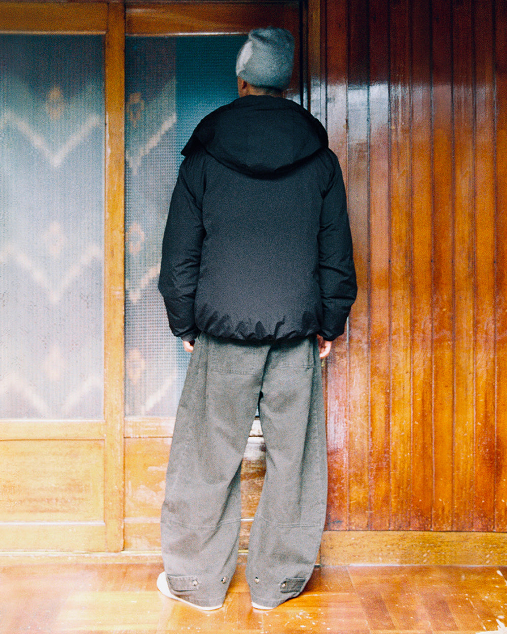 A person wearing a gray beanie, wide gray pants, and the Sundae School BLACK IBUL DOWN PUFFER with a detachable hood stands indoors facing a wooden wall with frosted glass panels.