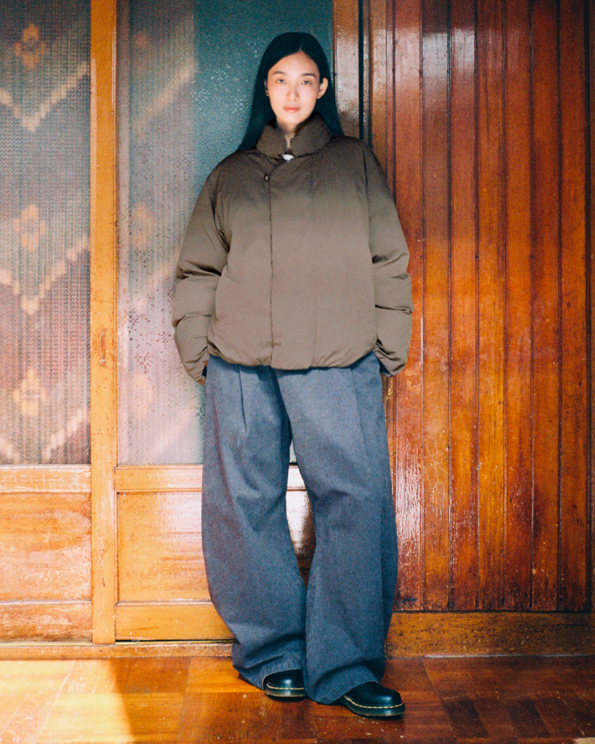 A person stands indoors wearing the Sundae School DEEP BROWN IBUL DOWN PUFFER with a detachable hood, paired with wide-leg blue trousers and black shoes. The background features wooden paneling and a glass door.