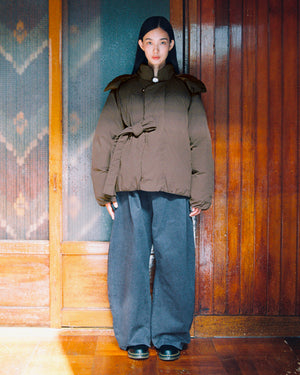 A person stands indoors, dressed in the Sundae School DEEP BROWN IBUL DOWN PUFFER, styled with a tie at the waist, wide gray trousers, and black shoes. The background features wooden panels and a frosted glass door.