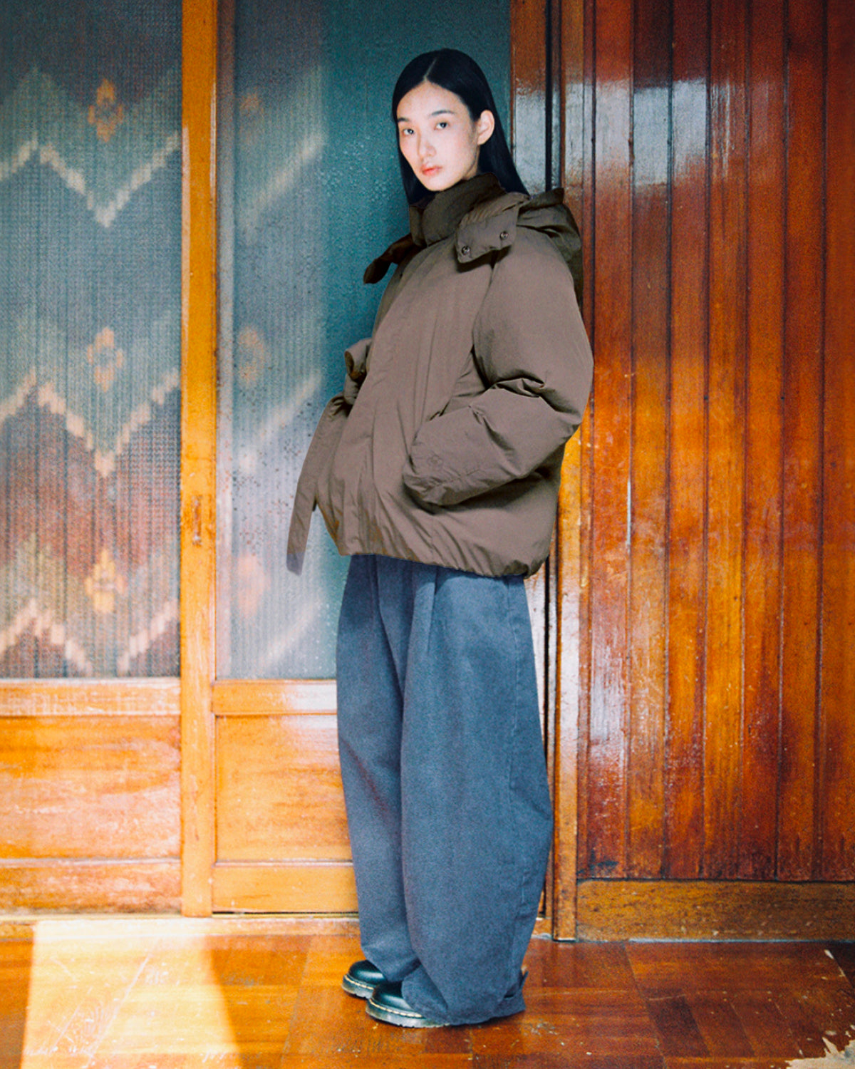 A person stands indoors, facing forward with their hands in the pockets of the DEEP BROWN IBUL DOWN PUFFER by Sundae School. They have paired the oversized brown down jacket with wide-leg gray pants and black shoes. Behind them are wooden doors and patterned glass.