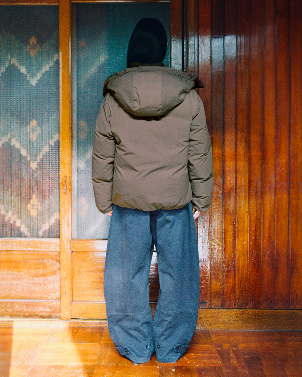 A person with long dark hair stands indoors facing wooden doors, wearing Sundae School’s DEEP BROWN IBUL DOWN PUFFER—an oversized down jacket with a detachable hood—paired with wide gray pants.
