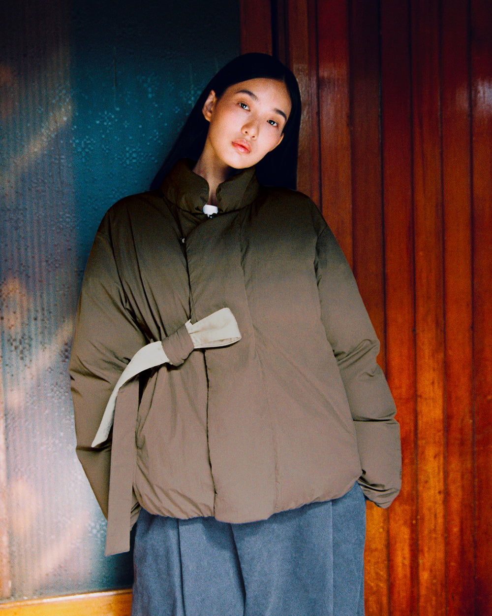 A person with long dark hair wears the Sundae School DEEP BROWN IBUL DOWN PUFFER, an oversized olive green jacket with a detachable hood and side tie, paired with loose gray pants, standing in front of wooden and glass panel doors.