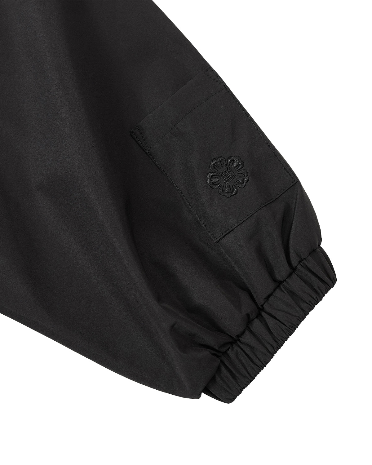 A close-up of the BLACK JEOGORI TIE SHELL JACKET by Sundae School highlights a black sleeve featuring an elastic cuff, a small pocket adorned with an embroidered floral design, and unisex technical details. Inspired by elements of the Korean jeogori jacket, this water-resistant outerwear piece blends traditional style with modern functionality.