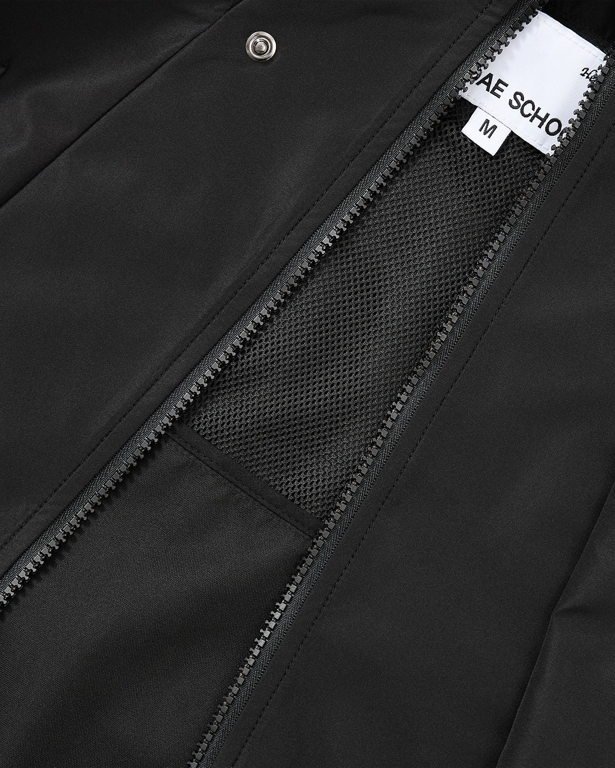 Close-up of the BLACK JEOGORI TIE SHELL JACKET by Sundae School, a unisex technical outerwear piece crafted from water-resistant materials. The partially open zipper reveals a mesh lining and a white clothing label marked M, with part of the brand name visible—showcasing the jacket’s seamless blend of style and functionality.