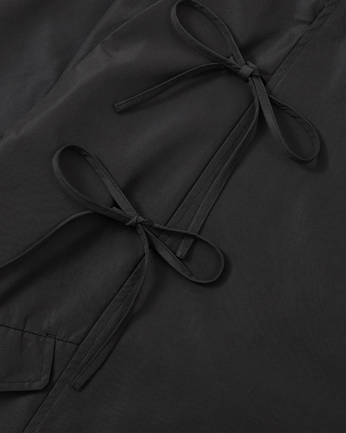 Close-up of the BLACK JEOGORI TIE SHELL JACKET by Sundae School, featuring black fabric with two neatly tied matching black bows, highlighting the unisex technical design’s clean stitching and smooth texture.
