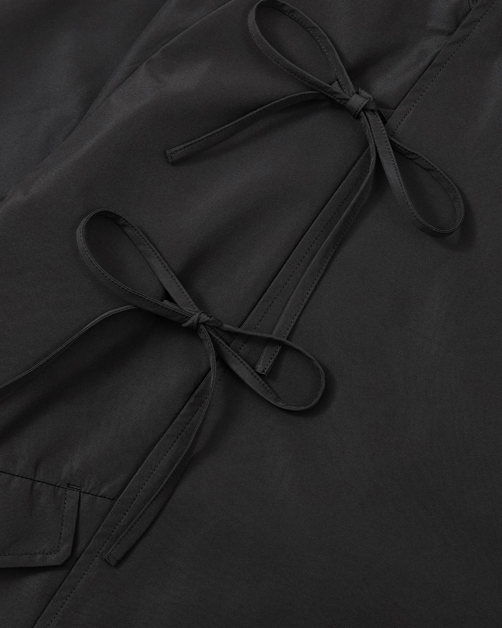 Close-up of the BLACK JEOGORI TIE SHELL JACKET by Sundae School, featuring black fabric with two neatly tied matching black bows, highlighting the unisex technical design’s clean stitching and smooth texture.