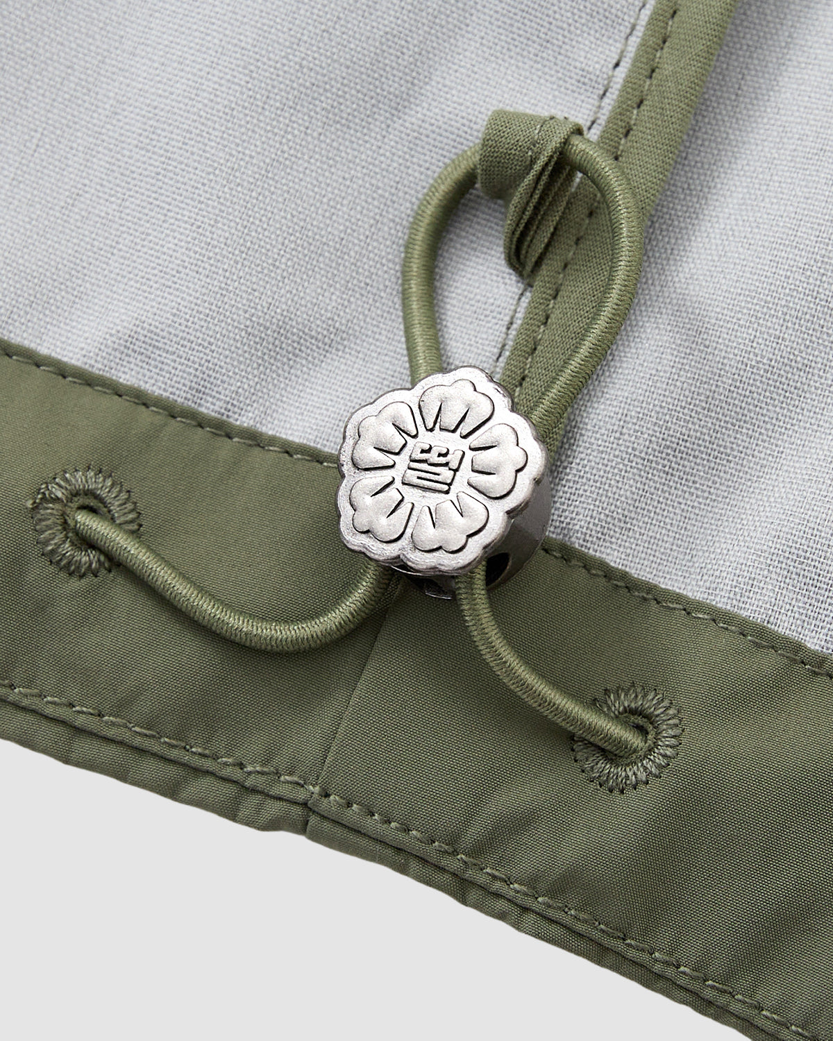 Close-up of a green drawstring with a silver flower-shaped cord lock, engraved symbol, and reinforced stitching on light gray and green fabric from the Sundae School MATCHA JEOGORI TIE SHELL JACKET, a water-resistant Korean-inspired piece.