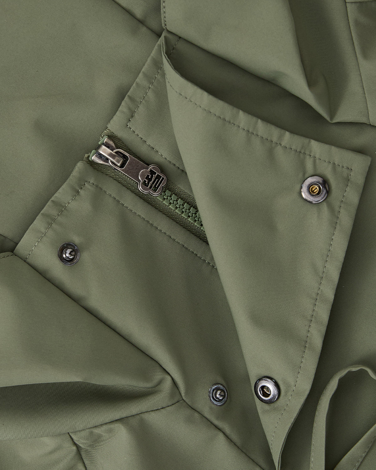 Close-up of the MATCHA JEOGORI TIE SHELL JACKET by Sundae School, a Korean-inspired olive green jacket with a zippered pocket, metallic snap buttons, and a smooth, slightly matte water-resistant finish.