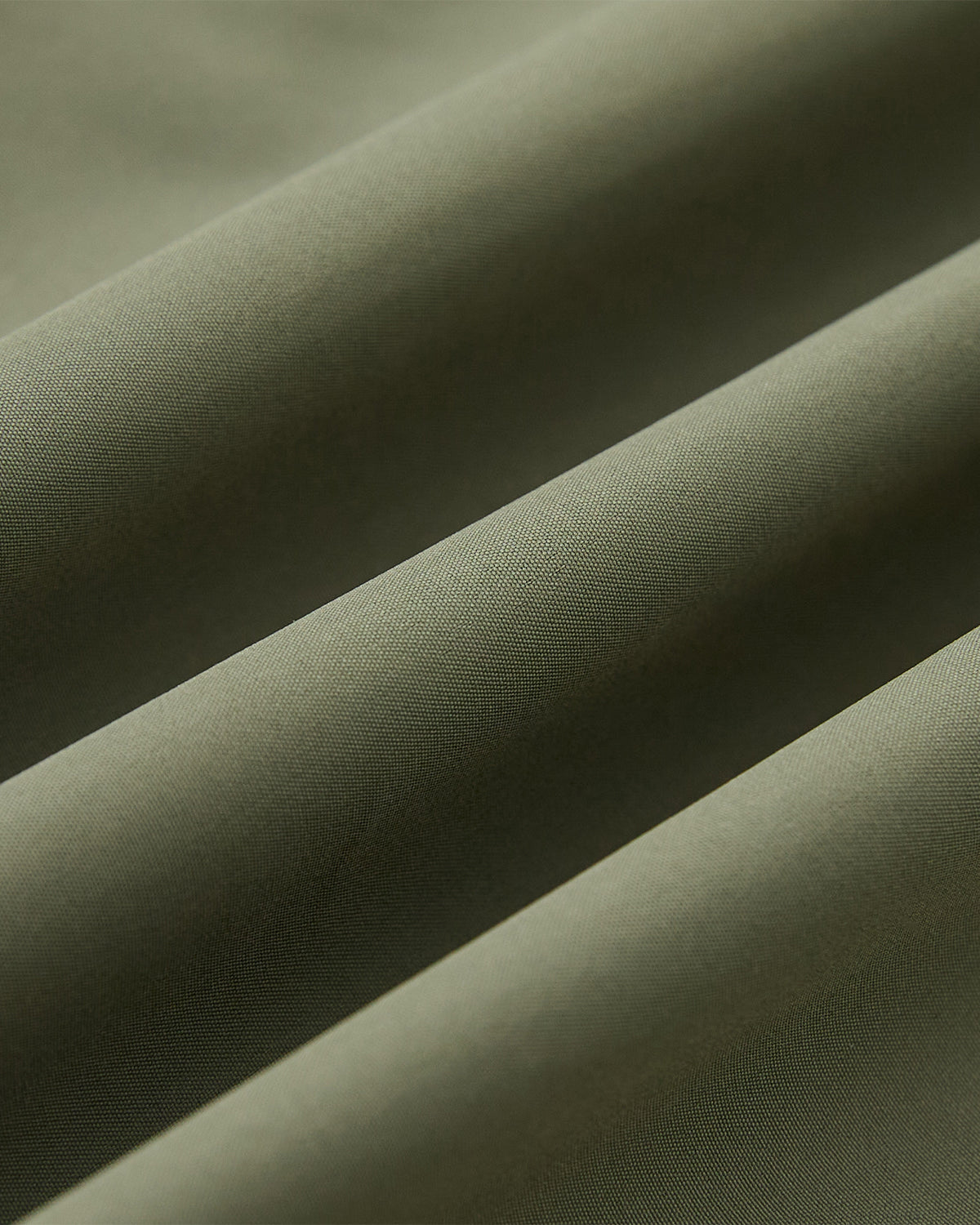 Close-up of smooth, olive green MATCHA JEOGORI TIE SHELL JACKET by Sundae School, with soft diagonal folds highlighting its water resistant texture and Korean-inspired drape.