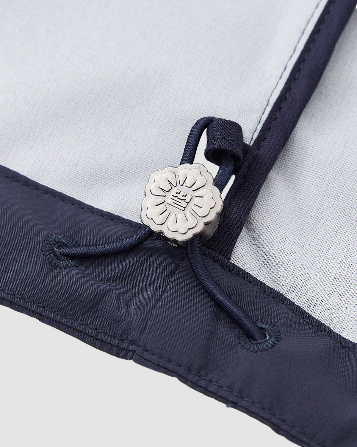Close-up of the Sundae School NAVY JEOGORI TIE SHELL JACKET: light gray and navy fabric with a dark blue drawstring, finished with a silver flower-shaped stopper. This water-resistant shell is inspired by the traditional Korean jeogori jacket.