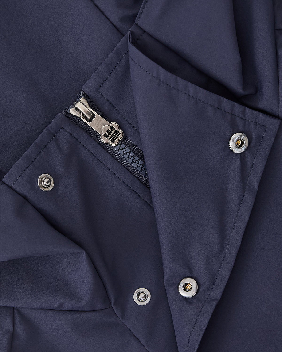 Close-up of the Sundae School NAVY JEOGORI TIE SHELL JACKET, a unisex technical outerwear piece in navy blue with metal snap buttons, a zipper featuring a 3D-engraved pull, and smooth, slightly shiny water-resistant fabric.