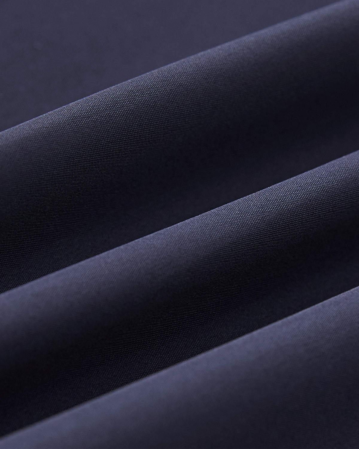 Close-up of the NAVY JEOGORI TIE SHELL JACKET by Sundae School shows folded dark blue, matte fabric. Soft diagonal lines and shadows highlight the fine weave and water-resistant shell of this unisex technical outerwear.