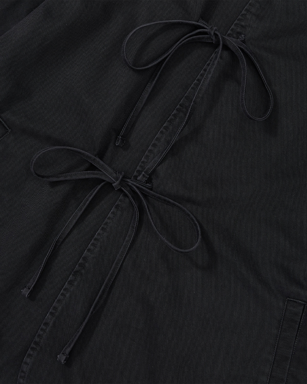 Close-up of the BLACK JEOGORI TIE JACKET by Sundae School in black fabric, highlighting two double goreum ties knotted in bows and set diagonally across the frame. Subtle stitching and pocket seams accent the textured textile, inspired by a monk collar jeogori silhouette and crafted from lightweight chino.