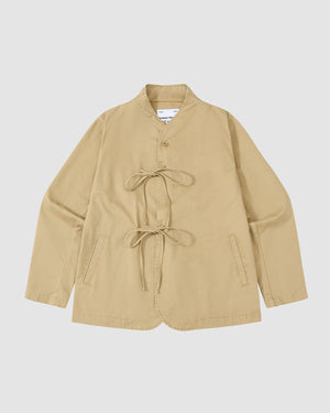 The TAN JEOGORI TIE JACKET by Sundae School features long sleeves, a monk collar, two front pockets, and front ties for closure in a classic jeogori silhouette. Laid flat on a white background. Ideal for summer suiting.