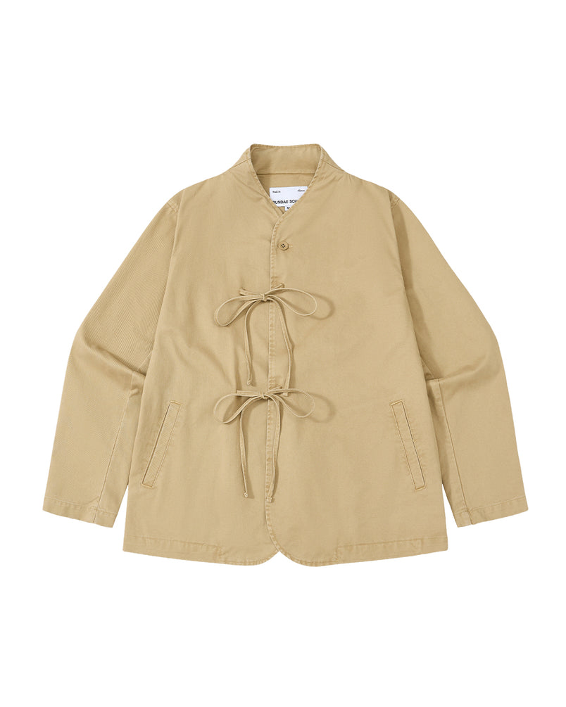 The TAN JEOGORI TIE JACKET by Sundae School is a beige, long-sleeve jacket inspired by traditional jeogori design, featuring a monk collar, two front pockets, and two matching fabric tie closures at the front. Crafted for summer suiting, it is displayed laid flat on a white background.