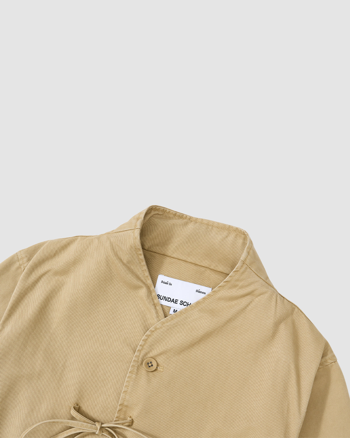 The Sundae School TAN JEOGORI TIE JACKET, a beige collared piece with button and tie closure inspired by the jeogori silhouette, is displayed flat on a white background. A white brand tag is visible inside the collar.