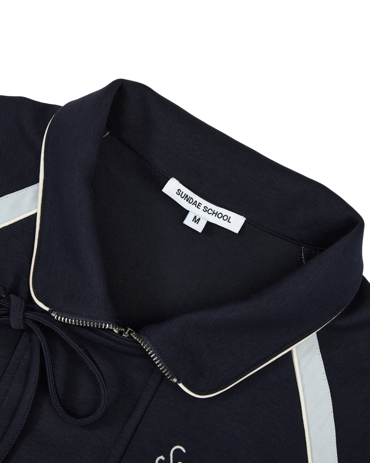 Close-up of the NAVY PALISADE PARK TRACK JACKET by Sundae School, an athletic track jacket in navy with white trim, featuring a collared design and a visible “SUNDAE SCHOOL M” clothing tag. Subtle chain-stitch embroidery offers refined detail to the classic silhouette.