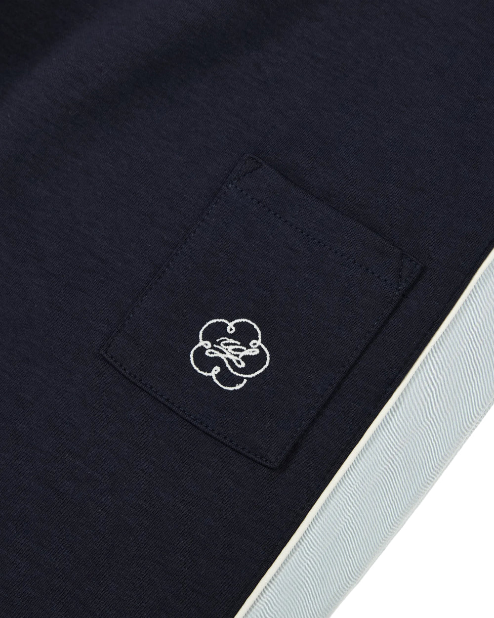 A close-up of the NAVY PALISADE PARK TRACK PANTS by Sundae School showcases the intricate white embroidered design and stitching on the navy blue pocket, set beside a crisp white stripe along the edge—highlighting the fine details characteristic of these breathable, stretch pants.