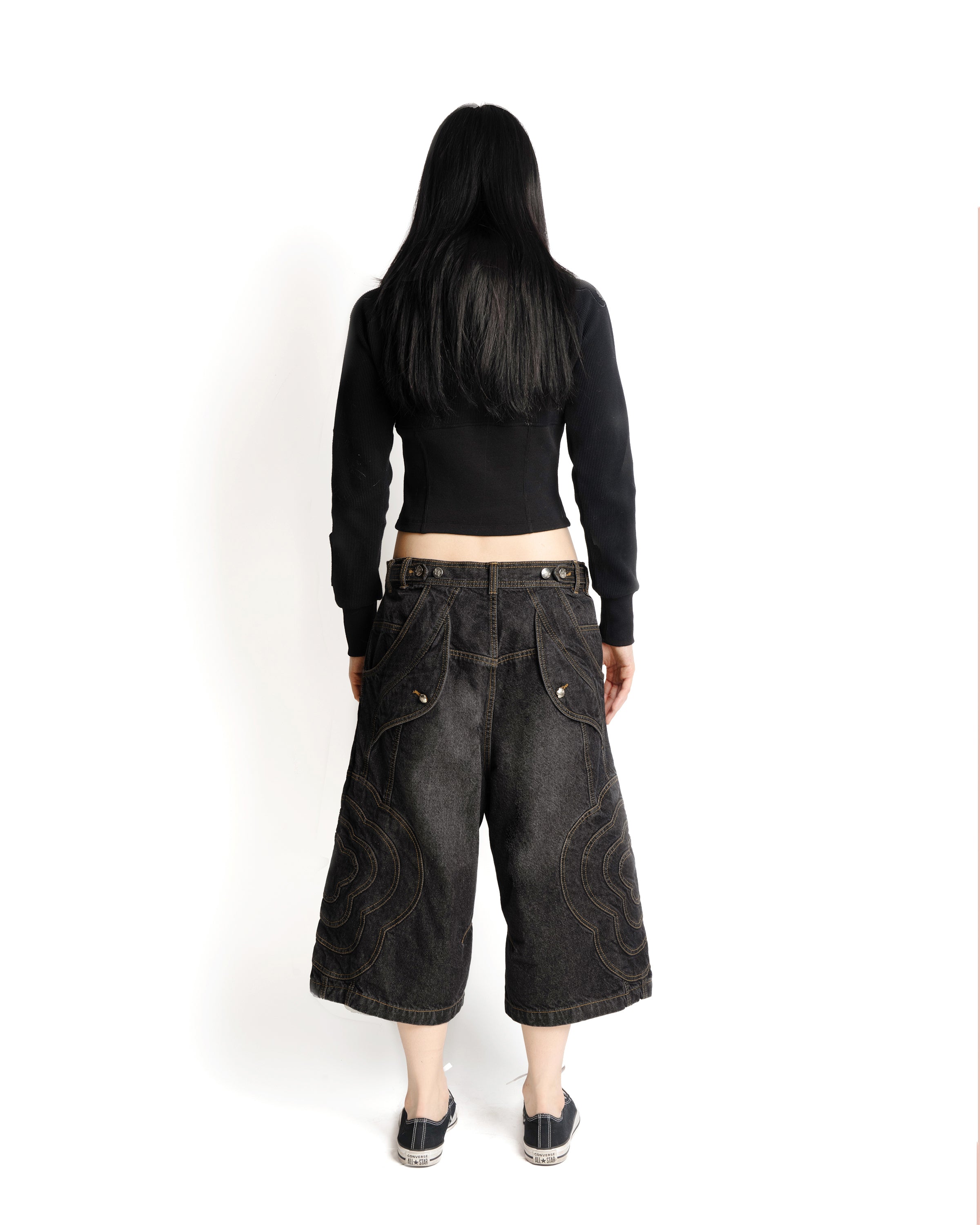A person with long black hair stands sideways against a white background, arms crossed, wearing Sundae School's BLACK JORTS JOONIVERSE—wide-leg dark denim jeans with a swirl pattern—paired with a black long-sleeve crop top and black sneakers.