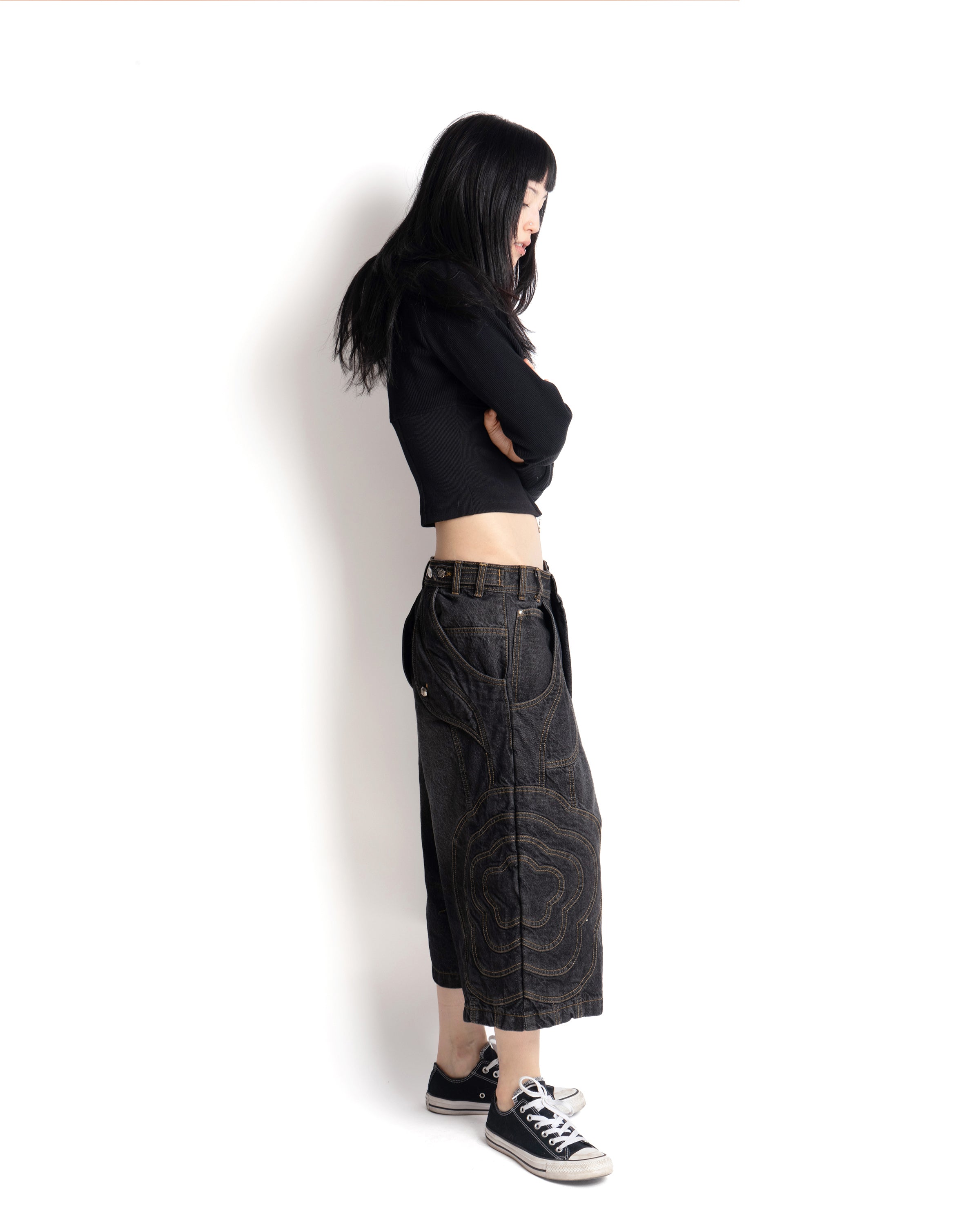 A person with long black hair, wearing Sundae School's BLACK JORTS JOONIVERSE and a black cropped jacket, paired with black slip-on shoes, stands facing away against a plain white background.