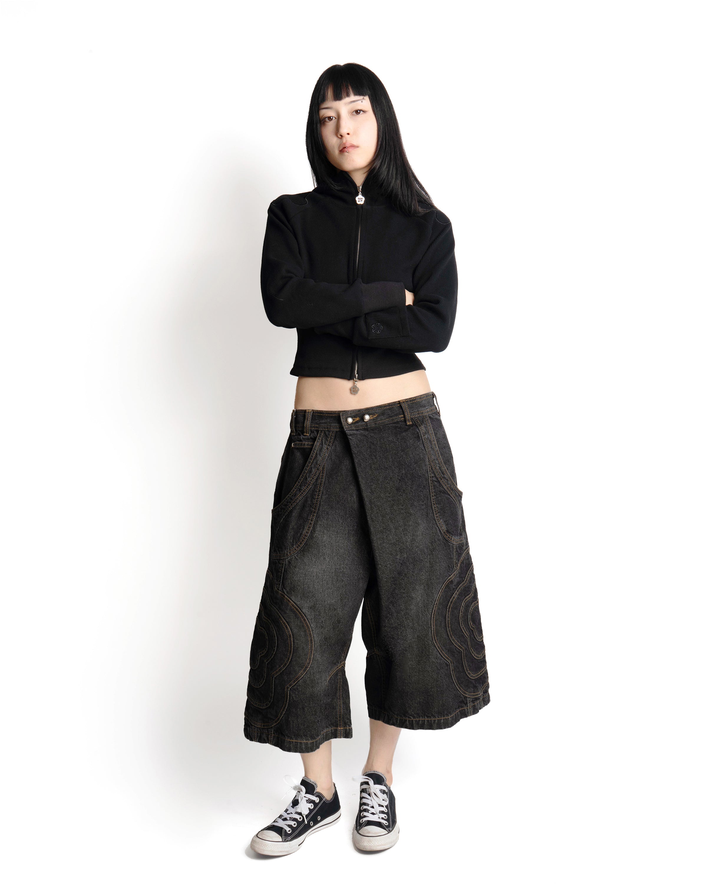 A person with long dark hair stands against a white background, arms crossed, wearing Sundae School’s BLACK JORTS JOONIVERSE—a black zip-up top, wide-leg dark denim shorts with decorative stitching—and black-and-white sneakers.