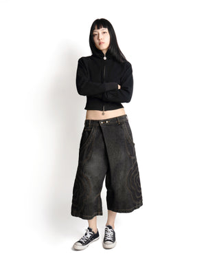 A person with long dark hair stands against a white background, arms crossed, wearing Sundae School’s BLACK JORTS JOONIVERSE—a black zip-up top, wide-leg dark denim shorts with decorative stitching—and black-and-white sneakers.