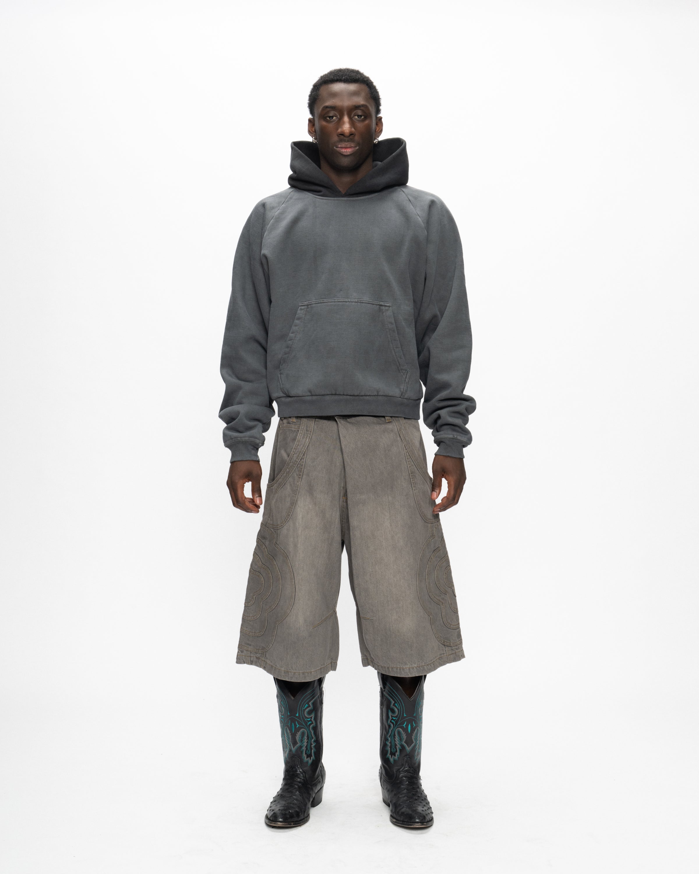 A person stands against a white background wearing Sundae School’s GREY JORTS JOONIVERSE and a dark gray hoodie, paired with black cowboy boots featuring turquoise details.