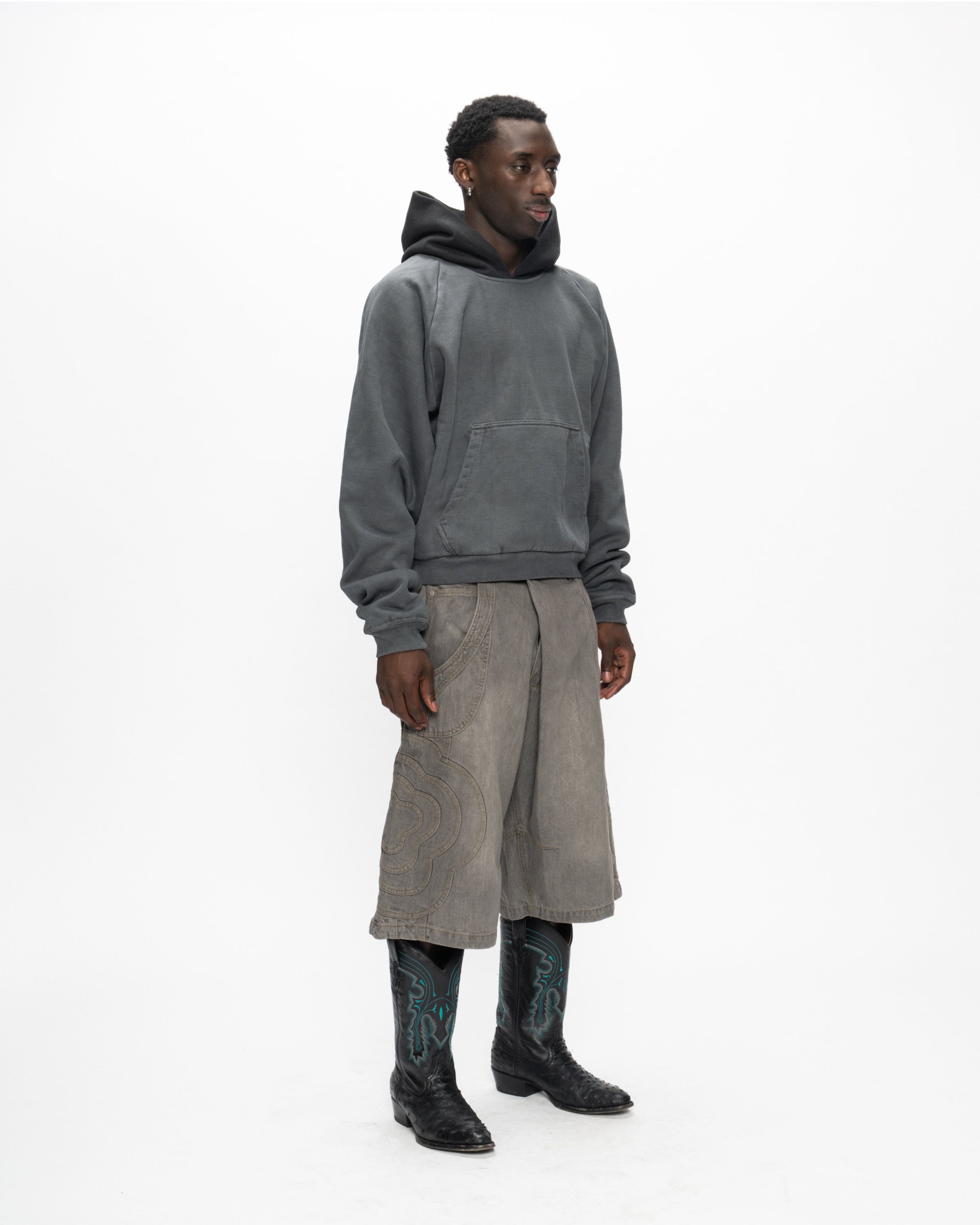 A person wears Sundae School's GREY JORTS JOONIVERSE—loose, patterned light gray shorts—paired with a dark gray hoodie and black cowboy boots with blue designs, standing against a white background facing slightly right.