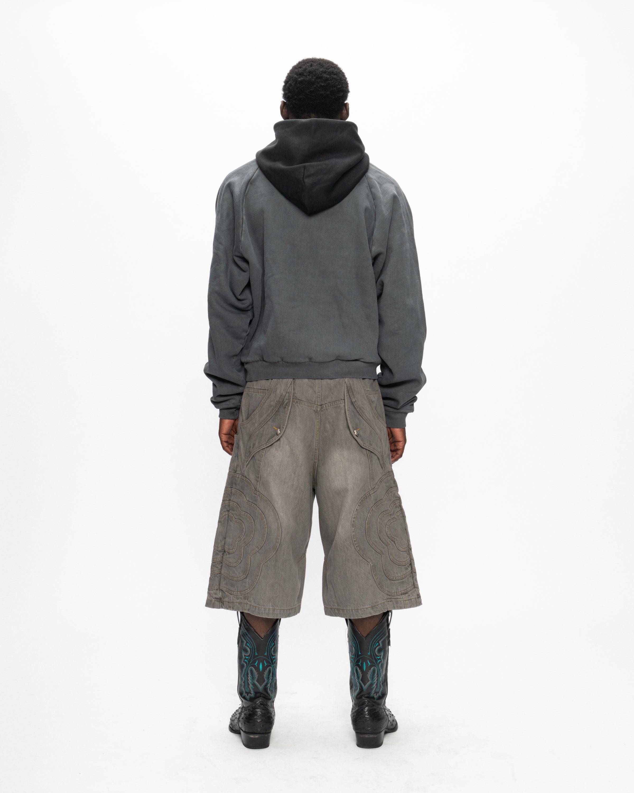 A person faces away on a white background, wearing Sundae School's GREY JORTS JOONIVERSE—oversized gray knee-length shorts—paired with a dark hoodie and black boots with blue patterns.