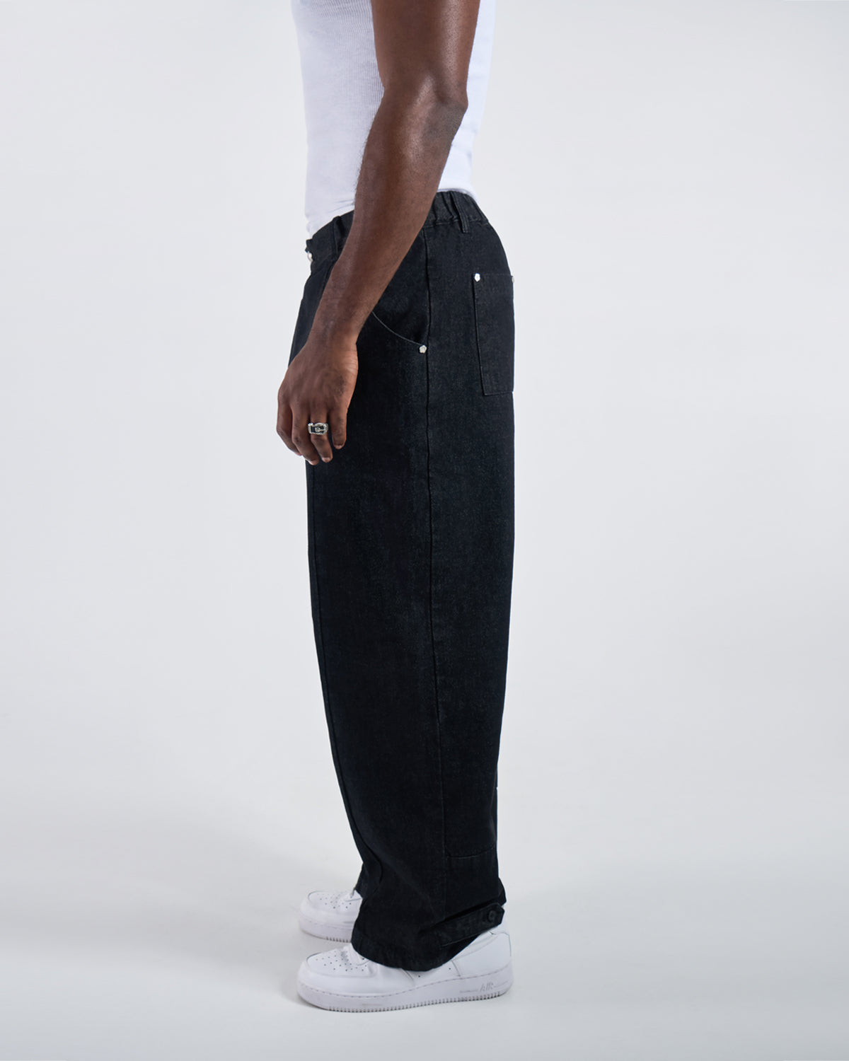 Side profile view of black wide-leg hanbok denim pants modeled with white ribbed tank top and silver ring, demonstrating relaxed fit silhouette and side seam details against white studio background