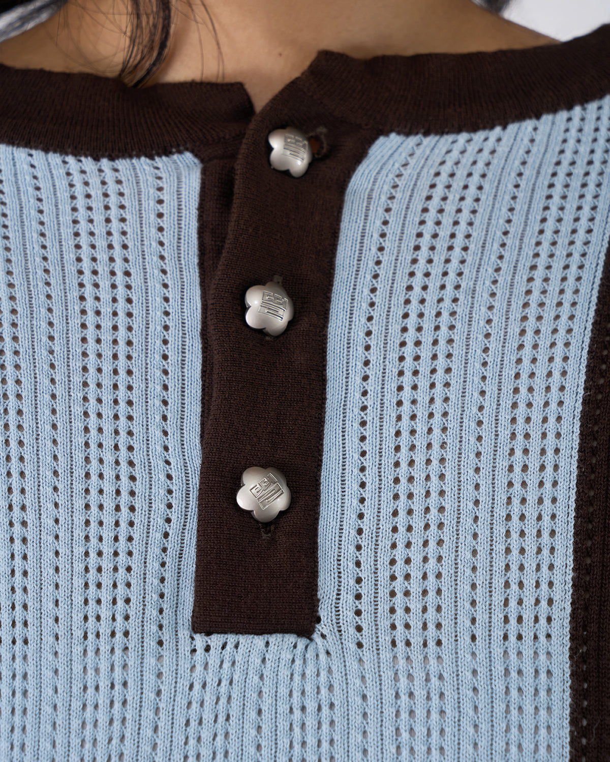 Close-up of Baby Blue Brown Sundae Soccer Jersey Knit henley button placket showing three white flower-shaped snap buttons with embossed details on brown knit placket, contrasted against light blue perforated mesh knit fabric, demonstrating construction and hardware quality.