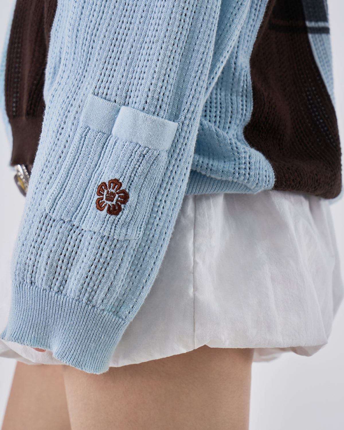 Close-up of Baby Blue Brown Sundae Soccer Jersey Knit light blue sleeve section showing small brown hibiscus flower embroidered detail on perforated mesh knit texture, demonstrating subtle branding elements and embroidery craftsmanship on garment extremities, styled with white mini skirt.