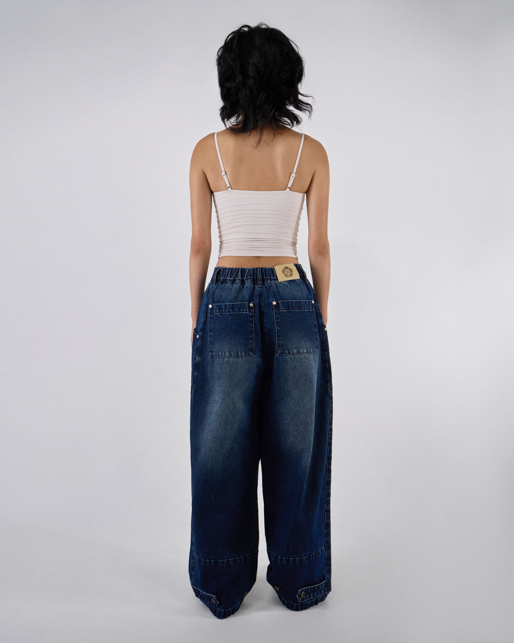 Back view of indigo fade wide-leg hanbok denim pants modeled with white ruched bandeau top, showing Sundae School leather patch, back pockets, and wide-leg silhouette against white studio background