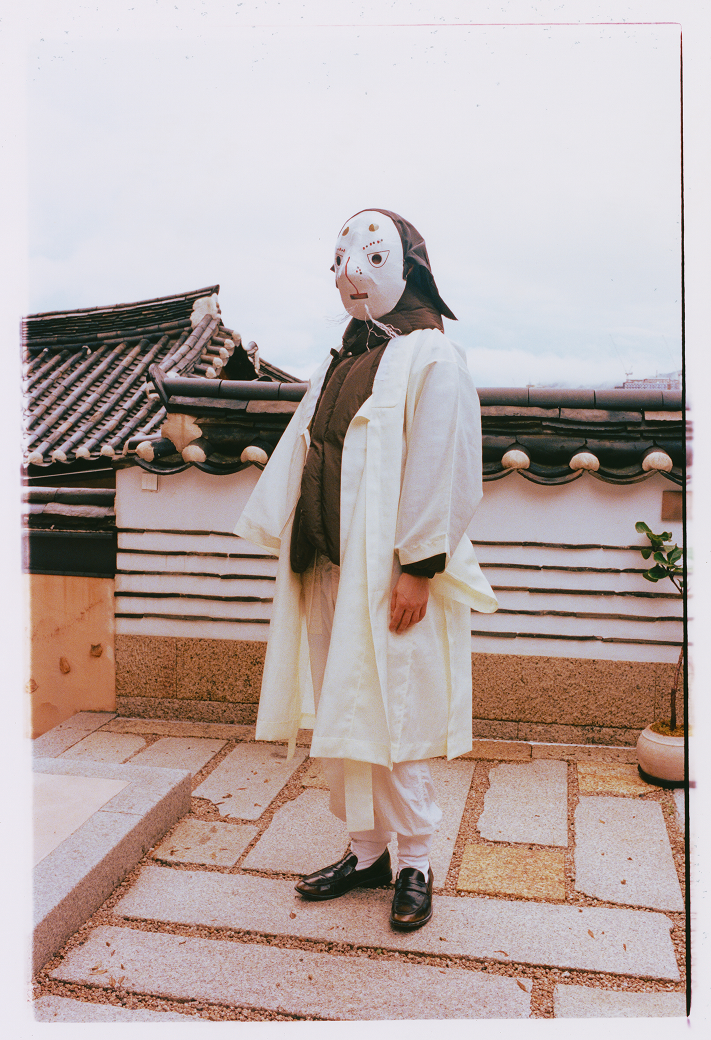 A male model wearing Sundae School Khaki Heavyweight Mochi Puffer in front of a Korean traditional house, Hanok.