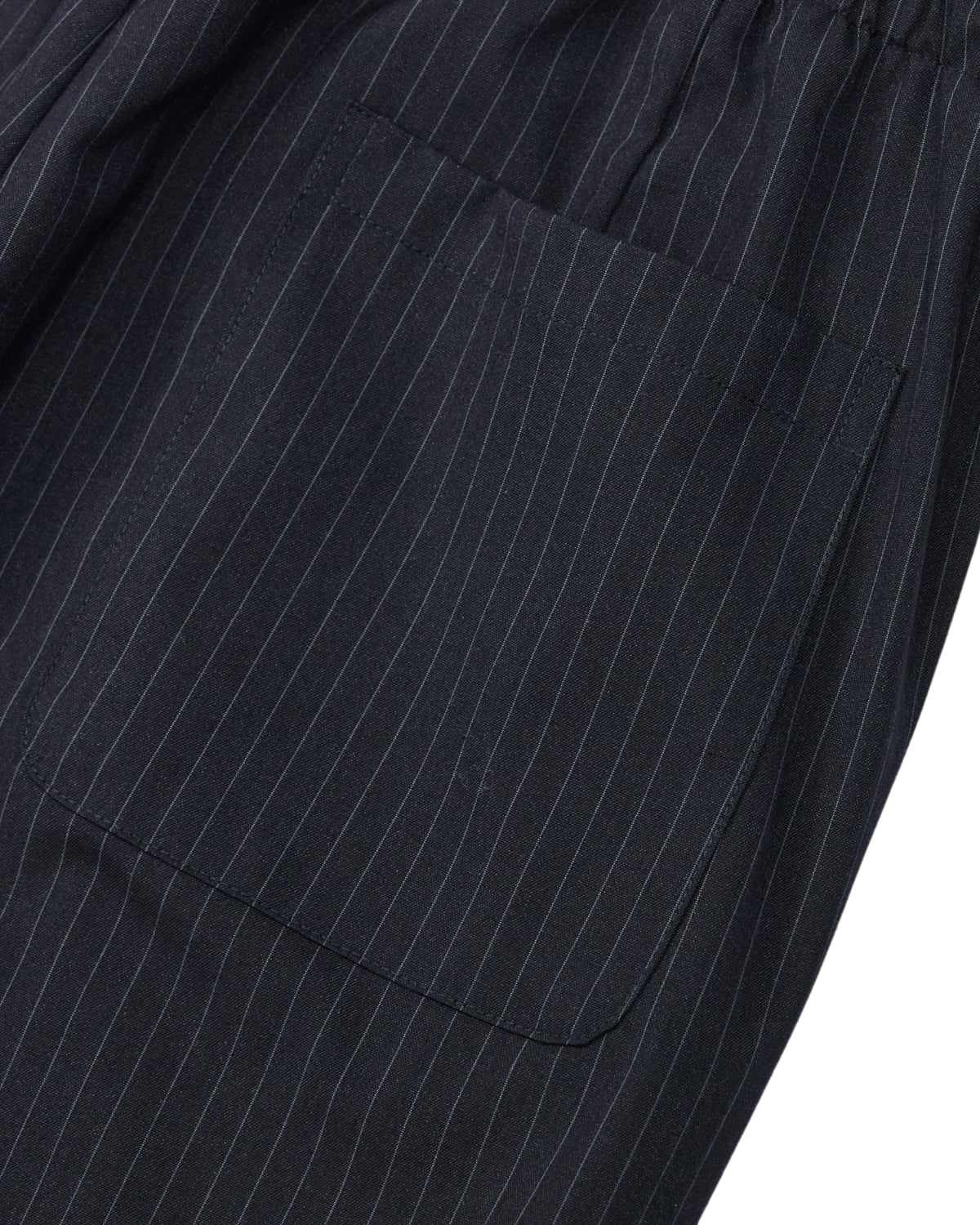 Close-up of the KIKI DRAWSTRING SLACKS by Sundae School: a dark fabric with thin vertical white pinstripes, showcasing a visible rectangular pocket sewn onto the material. The design evokes classic pinstripe trousers and features an elastic drawstring waist and a relaxed barrel leg silhouette.
