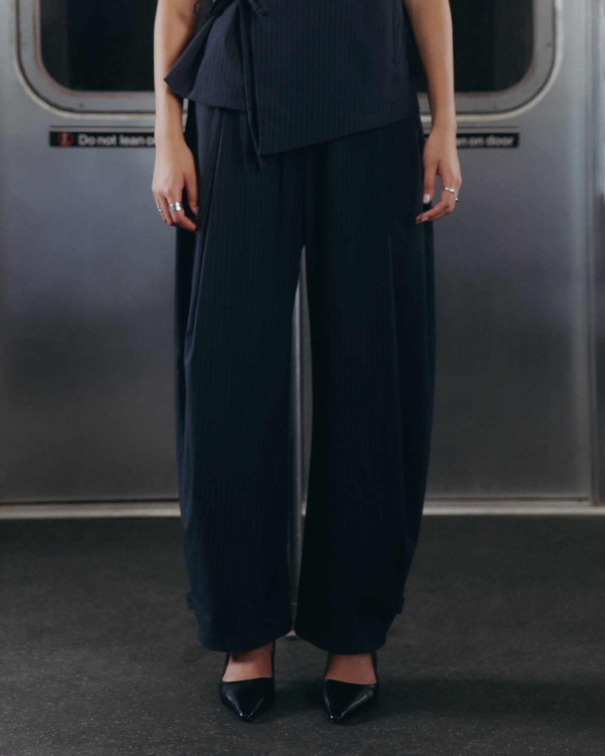 A person standing on a subway train floor, visible from shoulders down, wearing Sundae School’s KIKI DRAWSTRING SLACKS and black pointed-toe heels.