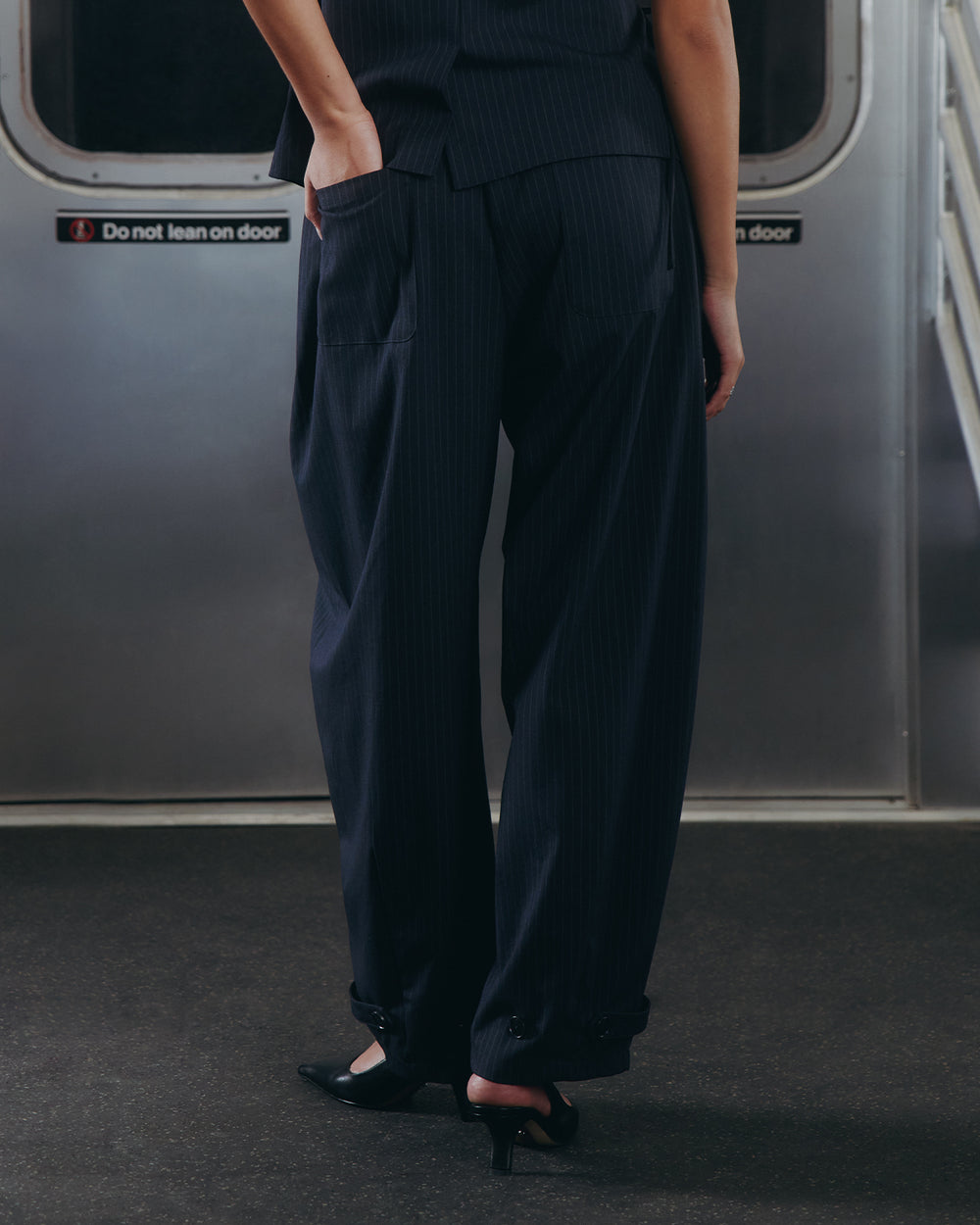 A person wearing Sundae School’s KIKI DRAWSTRING SLACKS stands in a subway car, facing away from the camera. The relaxed barrel leg pants are paired with black heels near a subway door displaying a Do not lean on door sign.