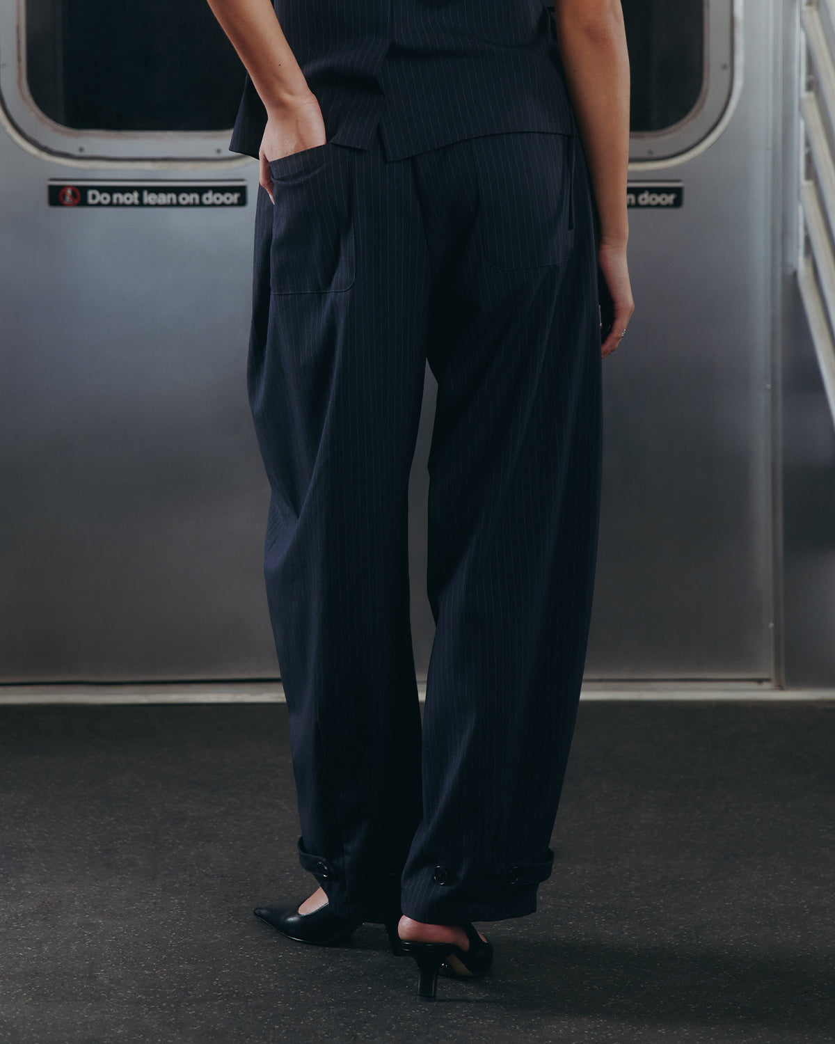 A person wearing Sundae School’s KIKI DRAWSTRING SLACKS stands in a subway car, facing away from the camera. The relaxed barrel leg pants are paired with black heels near a subway door displaying a Do not lean on door sign.