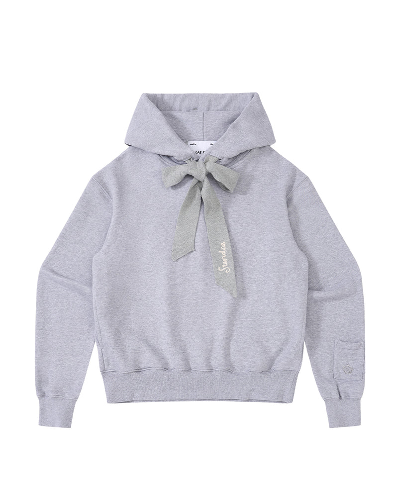 The HEATHER GREY KNIT TIE HOODIE by Sundae School is a light gray, heavyweight sweatshirt featuring a large knit bow at the neck, long sleeves, and ribbed cuffs and hem. Made from soft brushed cotton for extra comfort, this unisex hoodie is finished with “Salut” embroidered in cursive near the bow.