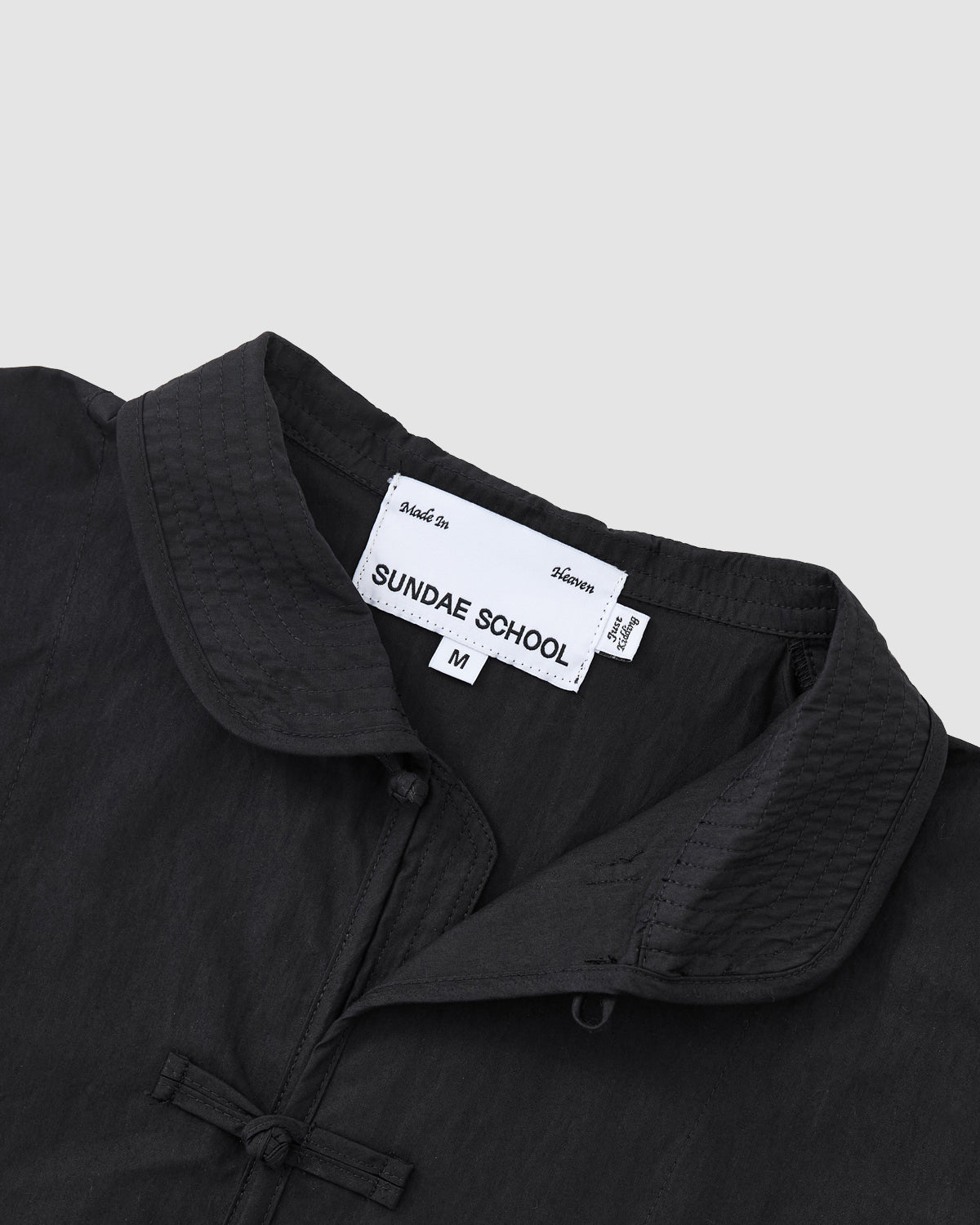 Close-up of the KNOT TIE TOWN JACKET by Sundae School, featuring a black textured collar and knotted button details inspired by Buddhist temple knots. The interior white tag displays 'Sundae School', size M, and 'Made in Korea.'.