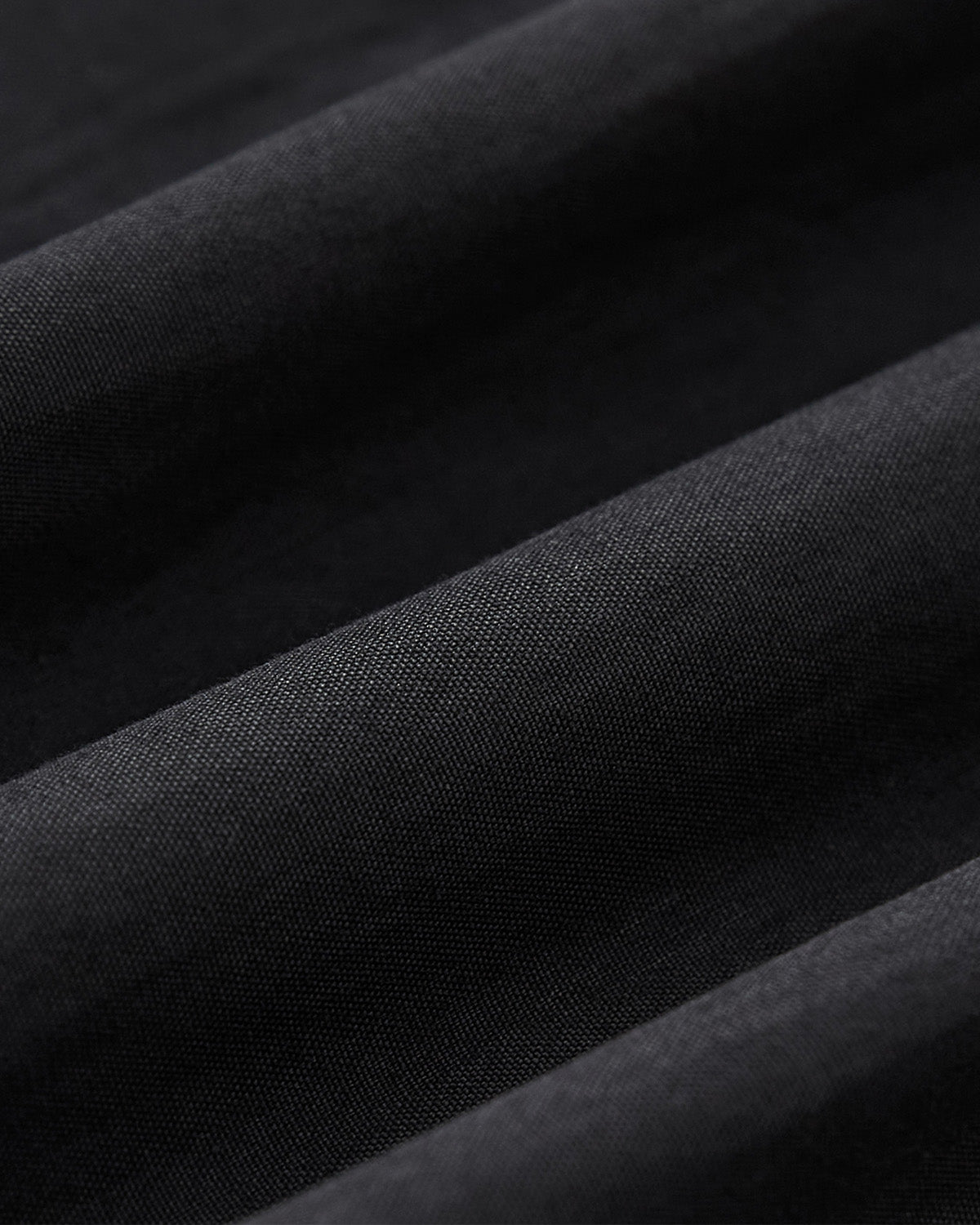 Close-up of black fabric with visible texture, gently folded into diagonal lines and soft shadows, reminiscent of patterns on the Sundae School KNOT TIE TOWN JACKET—an ideal unisex spring uniform layer.