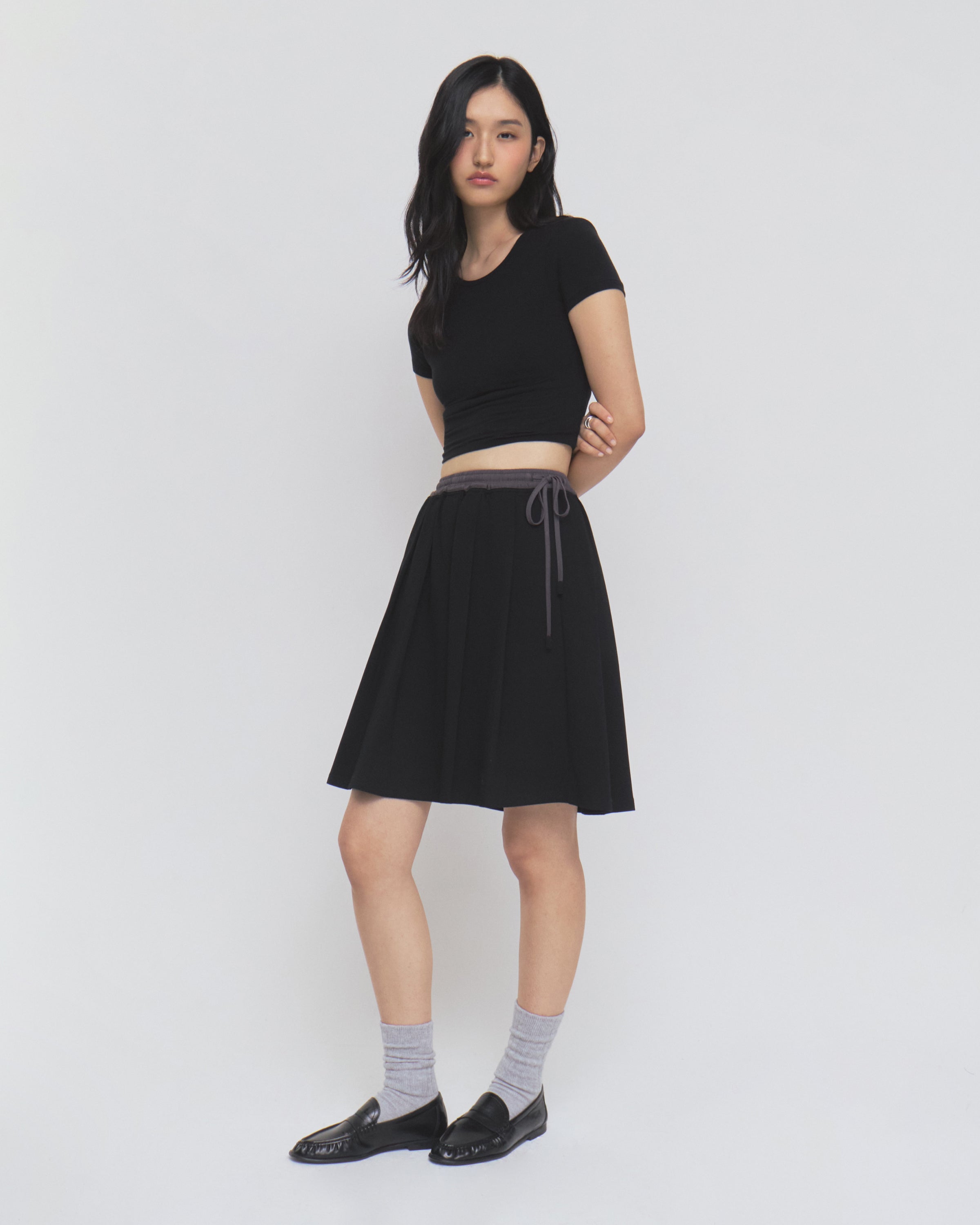 A woman with long dark hair and a neutral expression stands against a plain white background, wearing Sundae School’s BLACK KONA MIDI SKIRT paired with a black short-sleeve top, gray socks, and black loafers. The skirt features a gray drawstring detail.