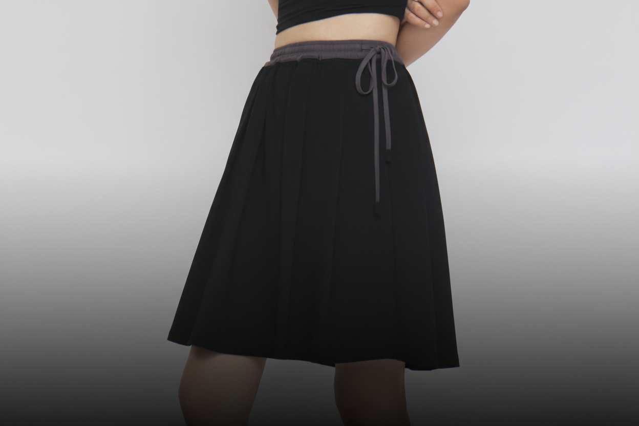 A woman with long dark hair and a neutral expression stands against a plain white background, wearing Sundae School’s BLACK KONA MIDI SKIRT paired with a black short-sleeve top, gray socks, and black loafers. The skirt features a gray drawstring detail. 