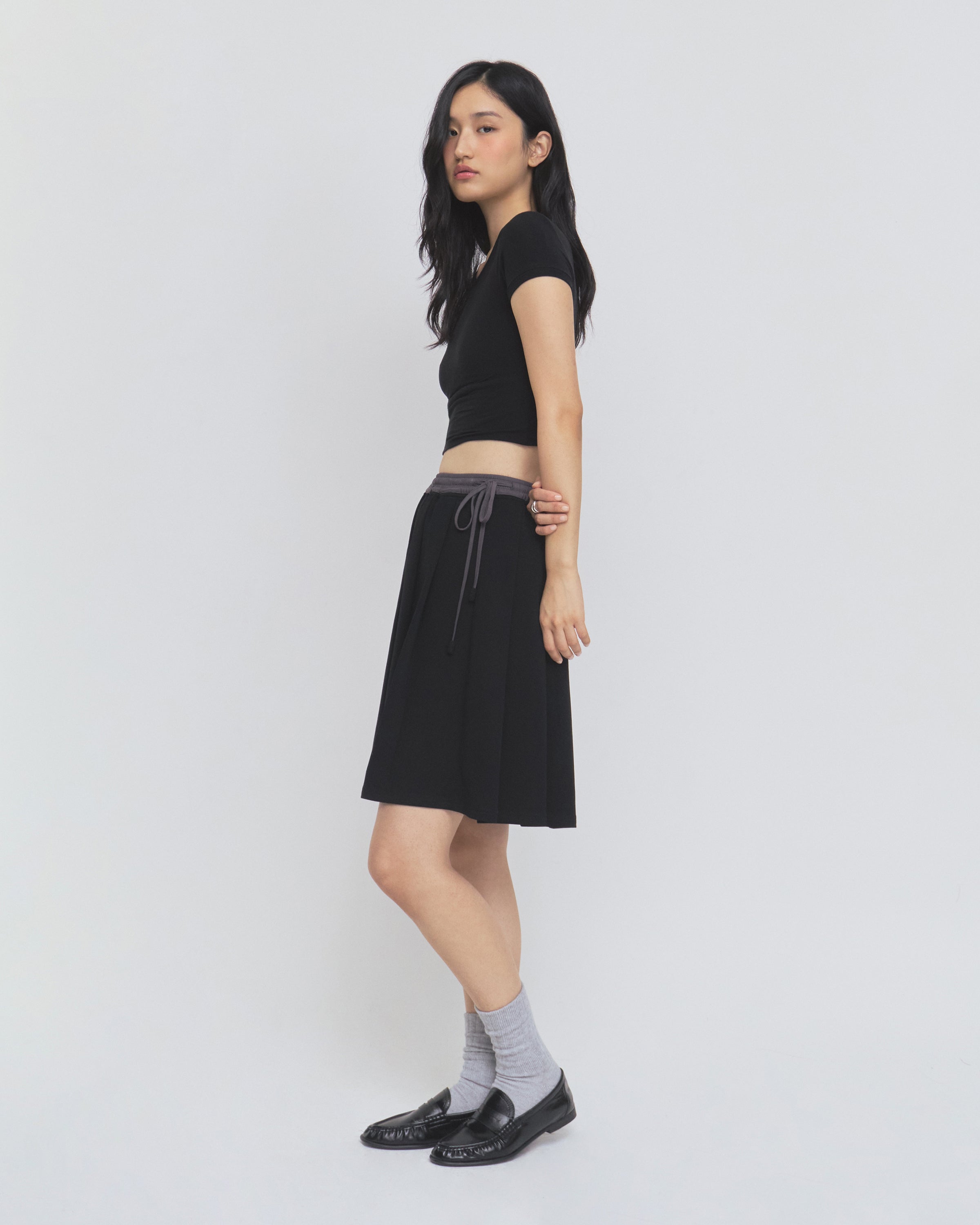 A woman stands sideways against a plain white background, wearing a black short-sleeve top, gray socks, black shoes, and the BLACK KONA MIDI SKIRT by Sundae School.