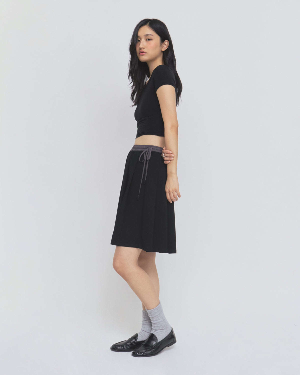 A woman stands sideways against a plain white background, wearing a black short-sleeve top, gray socks, black shoes, and the BLACK KONA MIDI SKIRT by Sundae School.