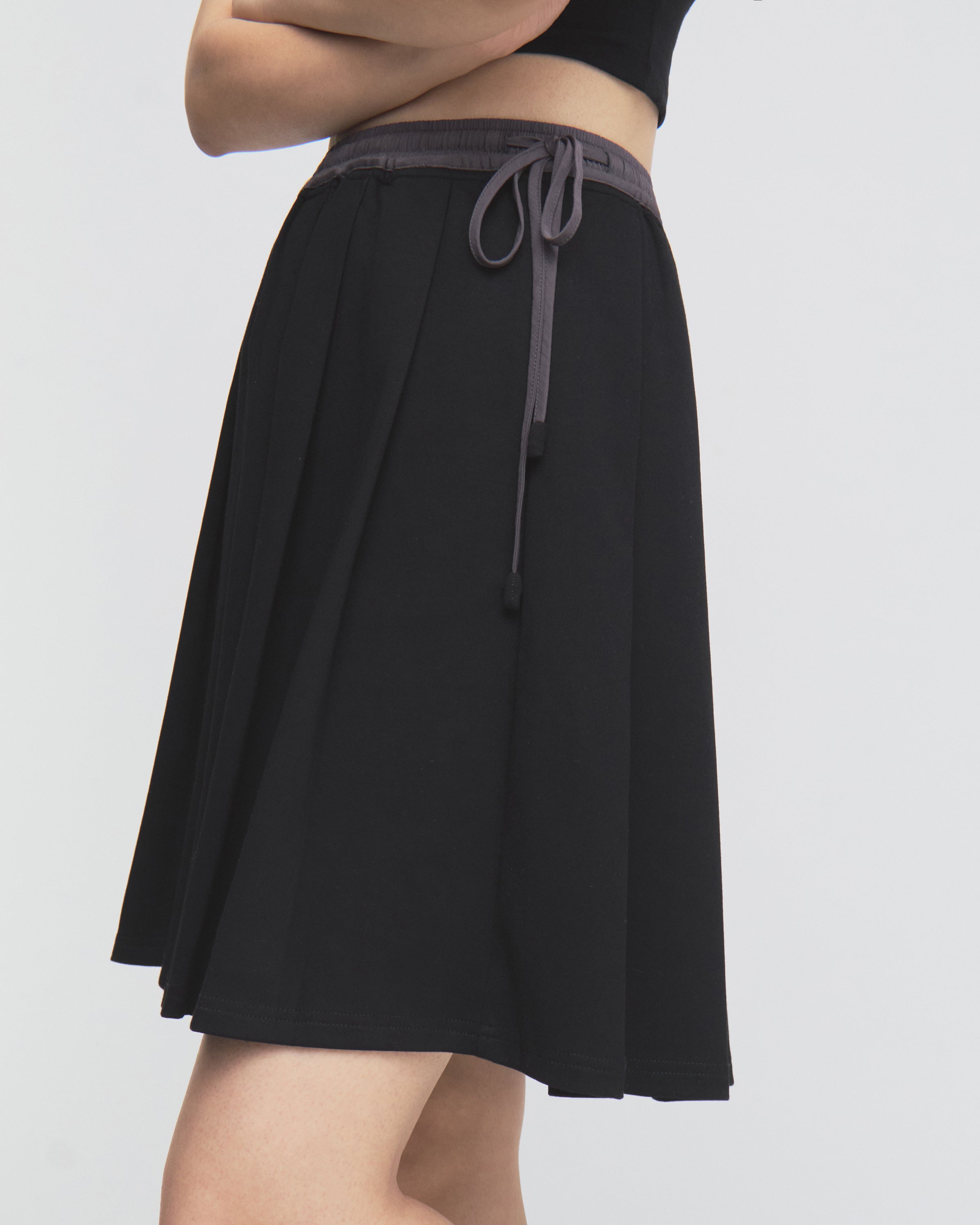 A model wearing the BLACK KONA MIDI SKIRT by Sundae School—a black skirt featuring a gray drawstring—paired with a black top, stands with arms crossed against a plain white background.