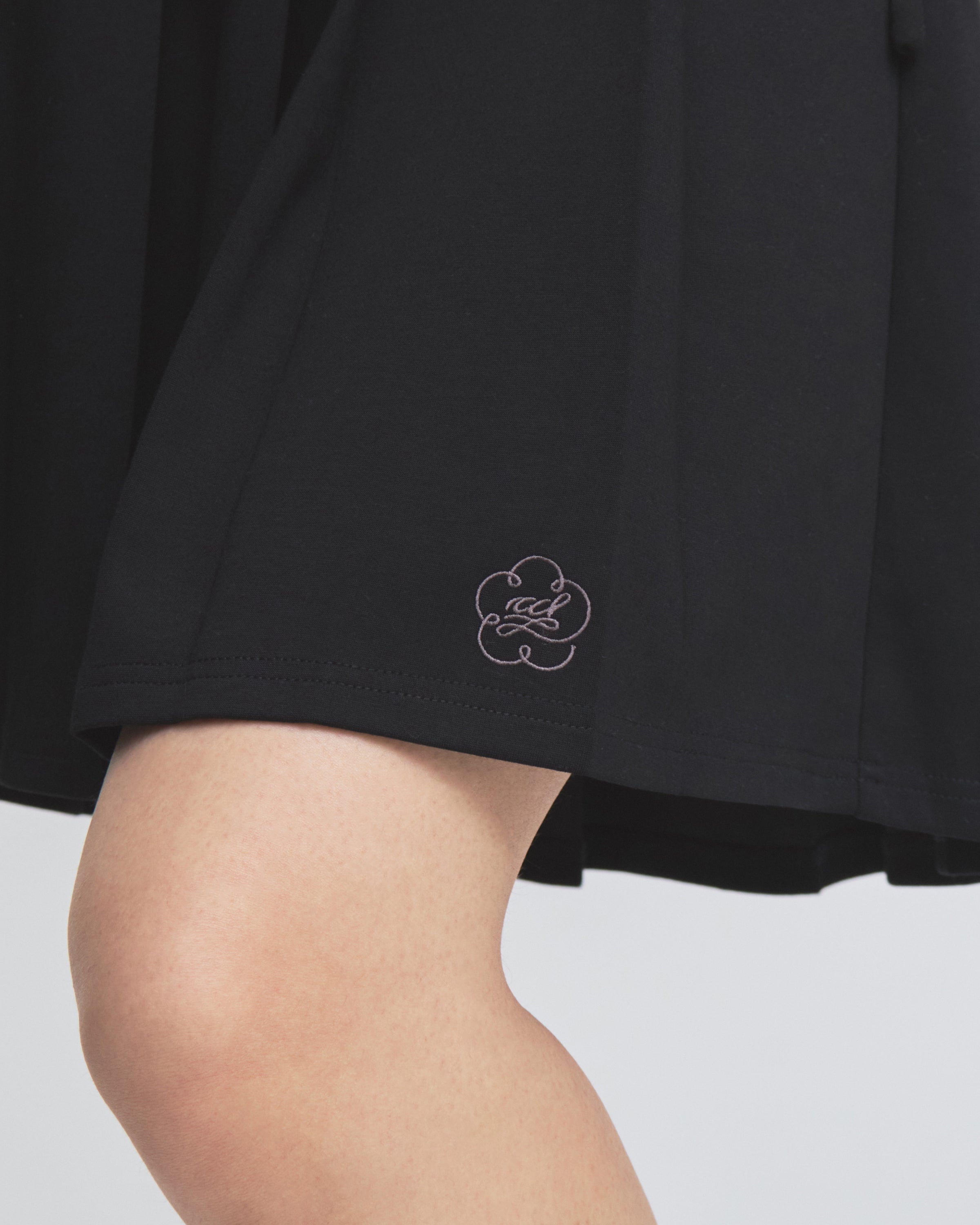 A close-up of a person’s knee and part of their upper leg, wearing the Sundae School BLACK KONA MIDI SKIRT, which features a small, pale flower-shaped logo near the hem. The background is plain and light-colored.