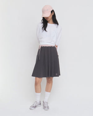 A person wearing a white long-sleeve shirt, the CHARCOAL KONA MIDI SKIRT by Sundae School, white socks, sneakers, and a light pink cap stands against a plain white background with their hands behind their back, looking down.